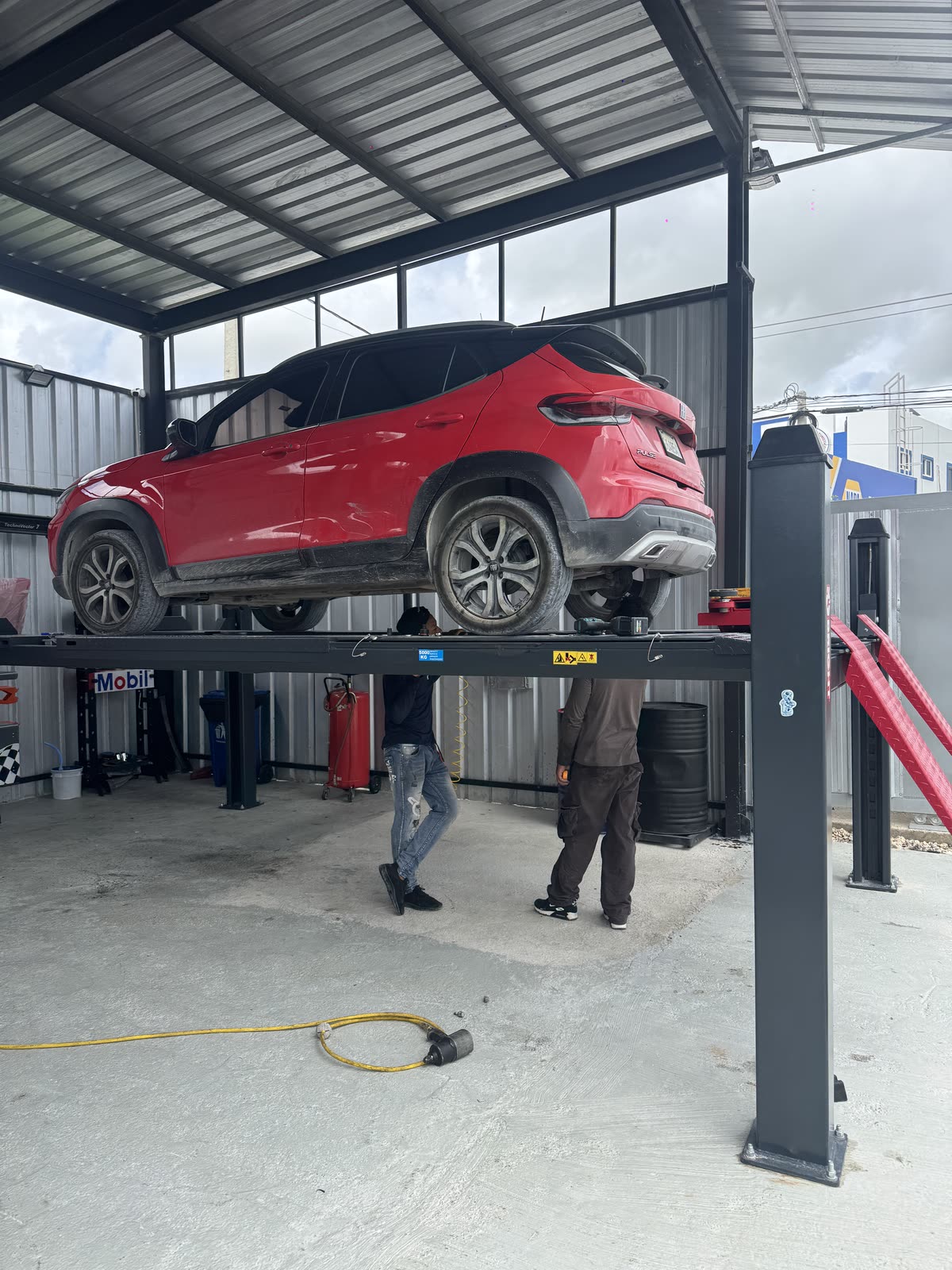 Carro rojo subido en el elevador de cuatro postes con dos mecánicos debajo